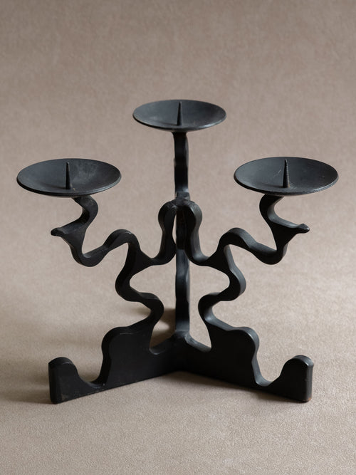 Closer front view of the forged iron three-arm candleholder, showing the pricket spikes, shallow dishes, curved arms, and black surface.