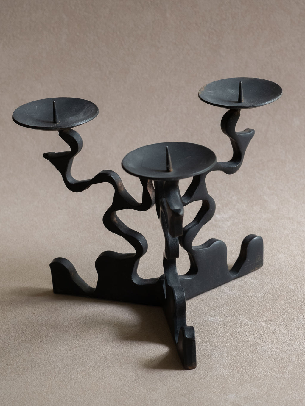 Three-arm forged iron candleholder shown from an angled front view, with blackened finish, raised pricket spikes, shallow candle dishes, and wavy sculptural arms.