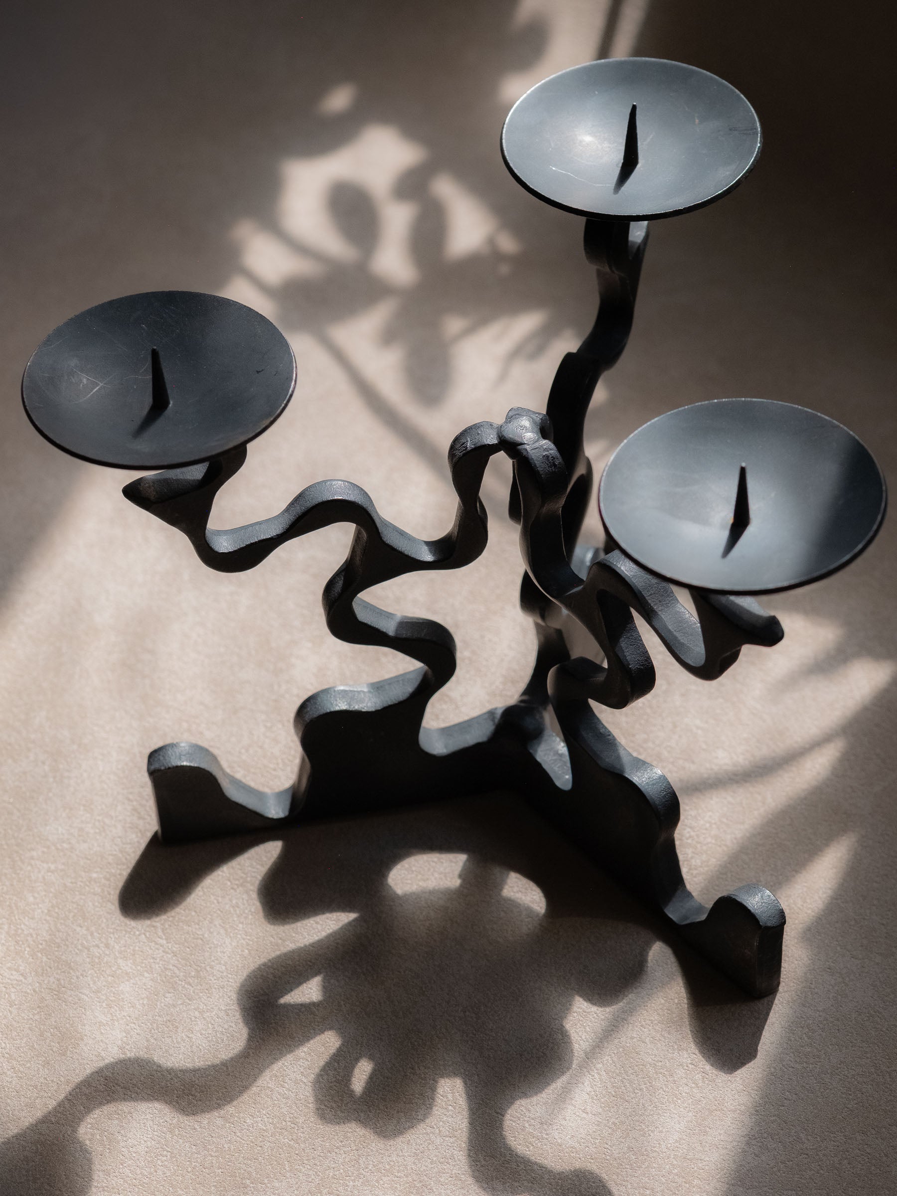 Top angled view of the three-arm forged iron candleholder, showing the three pricket candle dishes, sculptural arms, base, and cast shadows.
