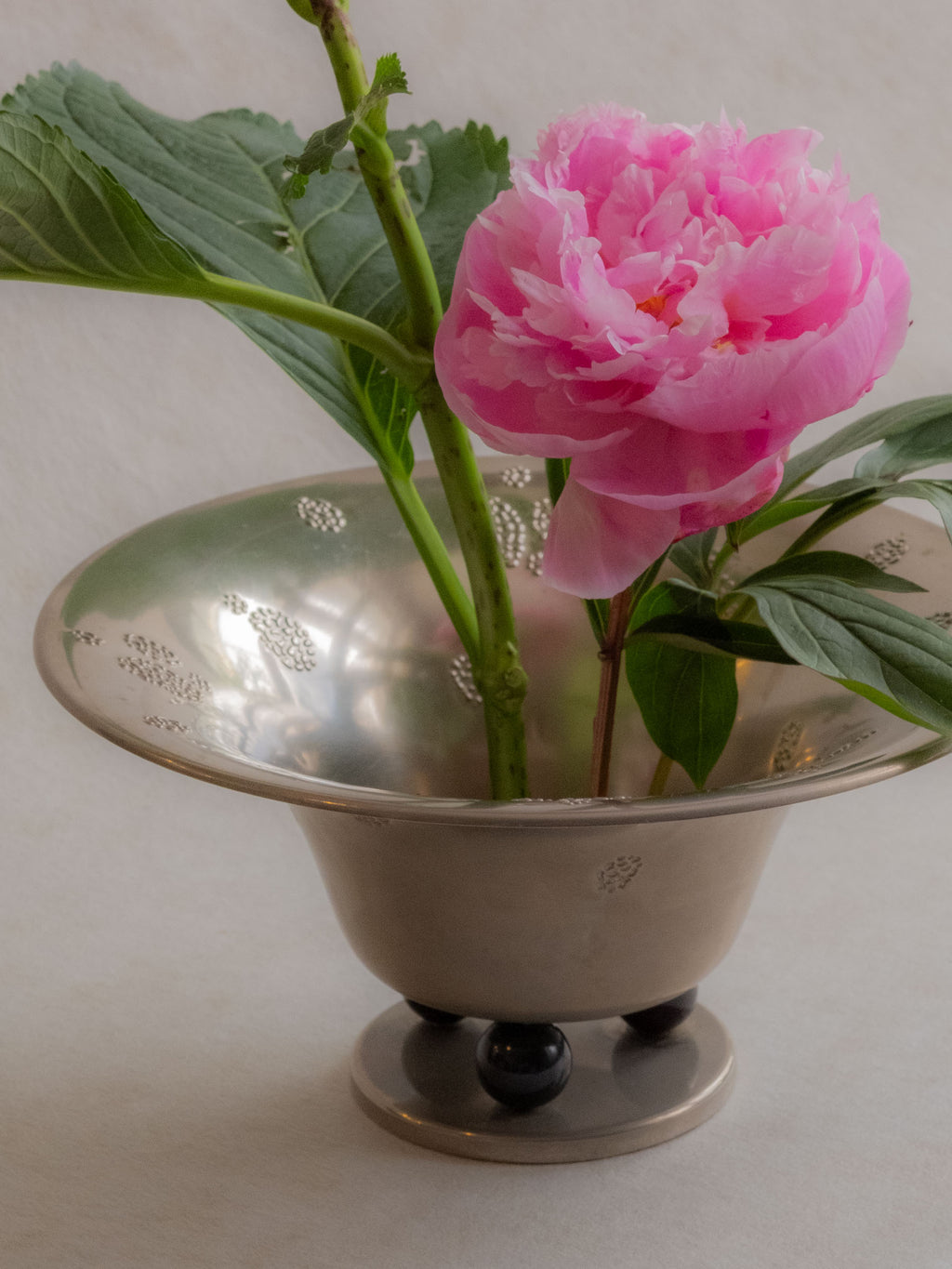 Close lifestyle view of a WMF metal pedestal tazza bowl arranged with pink flowers and green leaves.