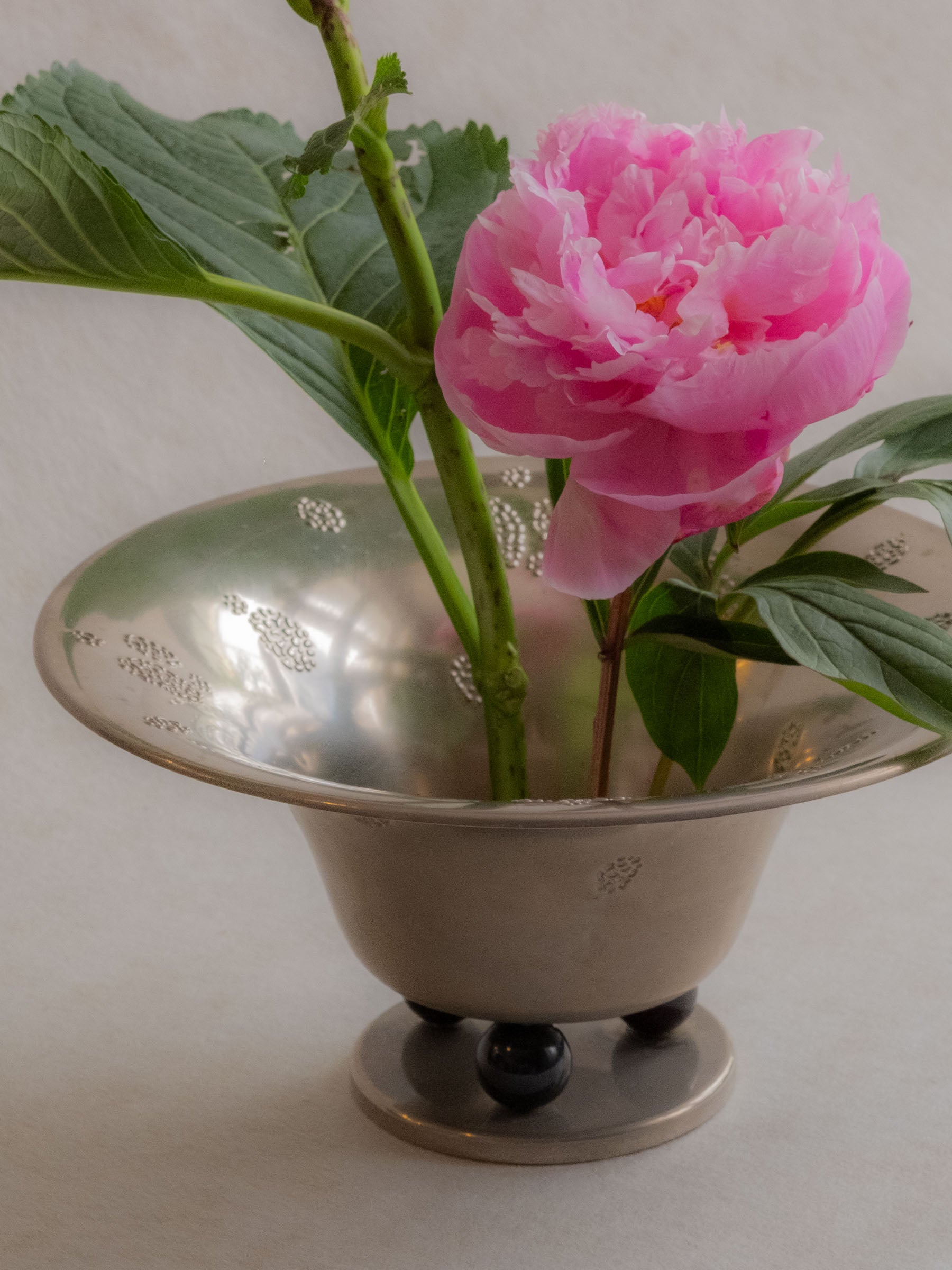 Close lifestyle view of a WMF metal pedestal tazza bowl arranged with pink flowers and green leaves.